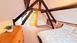 Ein Schlafzimmer mit schrägverdeckter Decke, freiliegenden Holzbalken, einem Bett mit gemusterten Betttextilien, einer hölzernen Kommode und einer Topfpflanze.