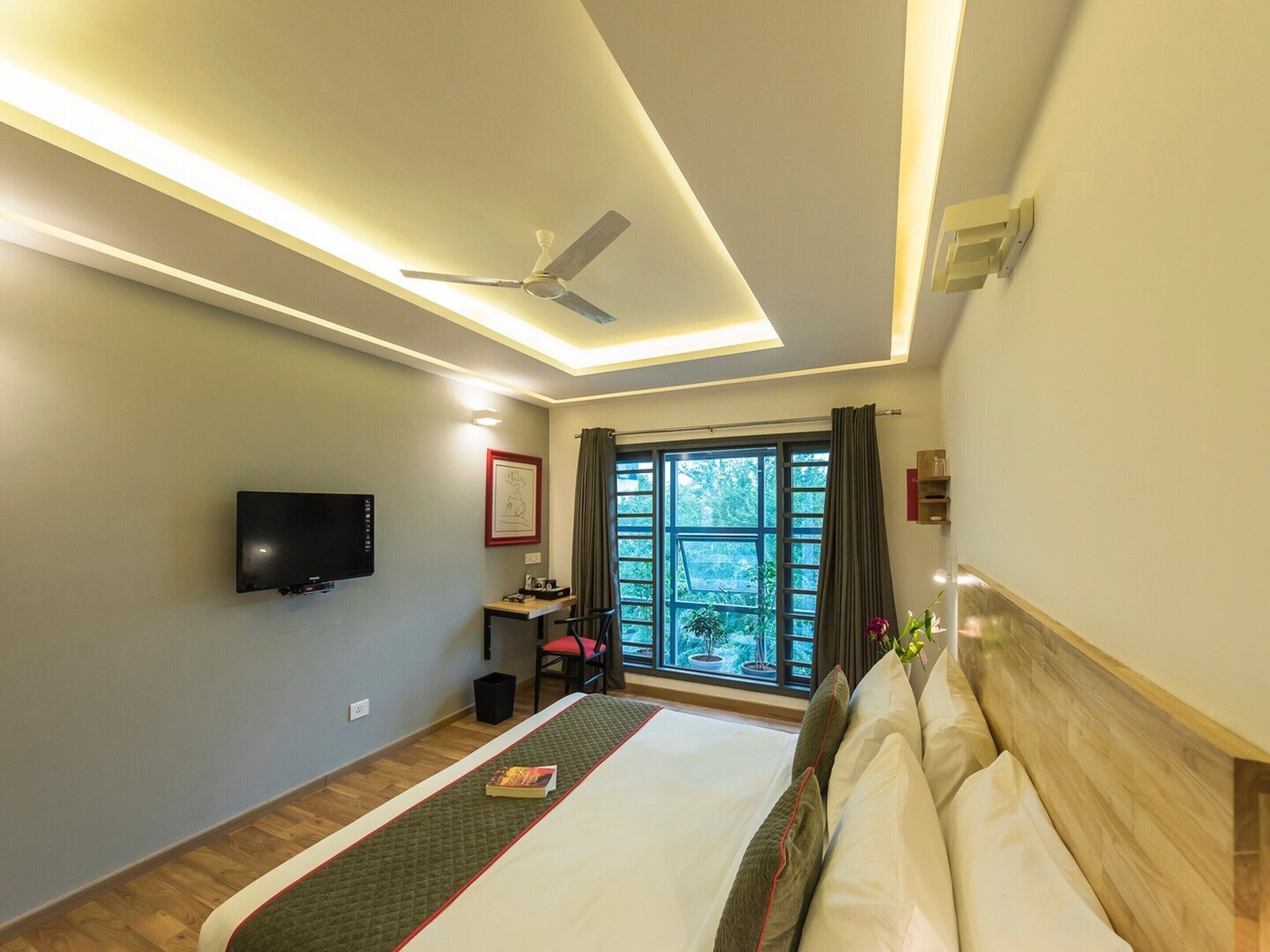 Une chambre d’hôtel équipée d’un lit, d’une télévision, d’un ventilateur de plafond, d’une fenêtre avec des rideaux et offrant une vue sur la verdure environnante.