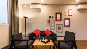 Una sala de estar moderna con un sofá, dos sillones, una mesa de centro y una lámpara de pie. Hay unidades de aire acondicionado en las paredes y cuadros enmarcados.