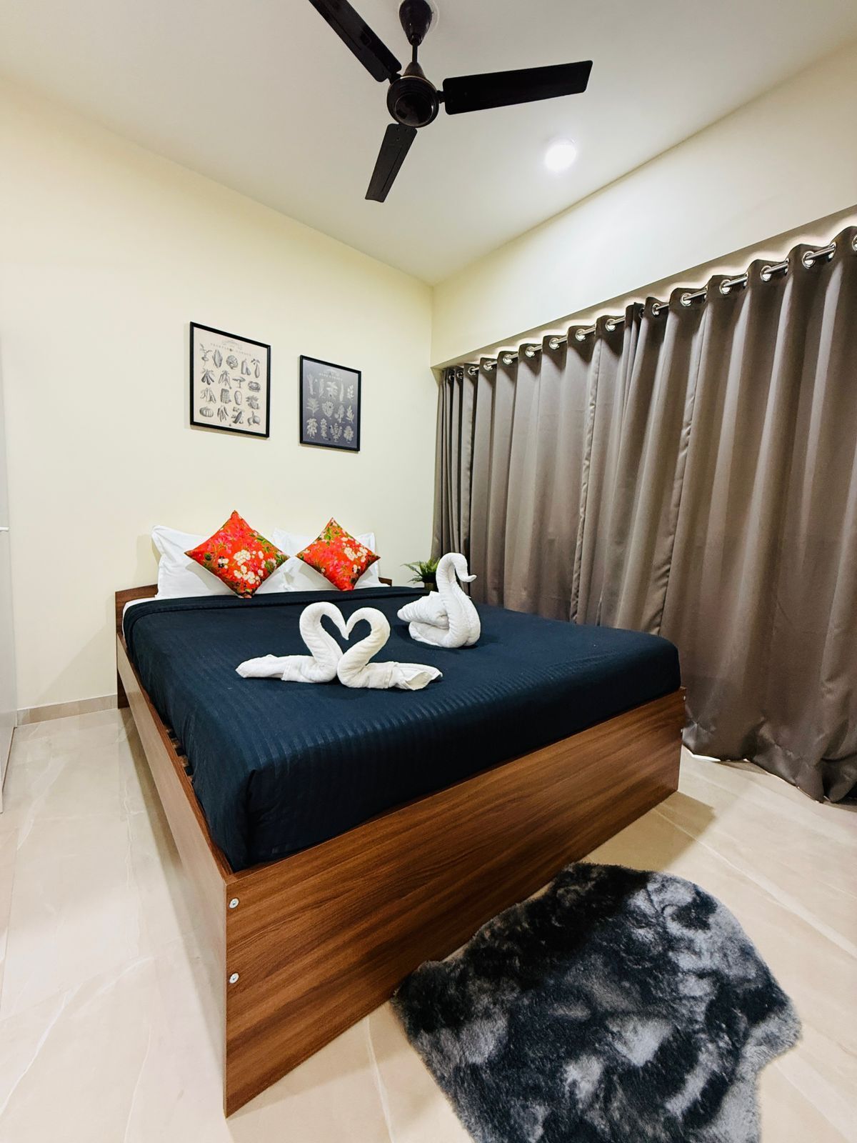 A bedroom with a bed, a ceiling fan, two framed pictures, a window with curtains, and a black rug.