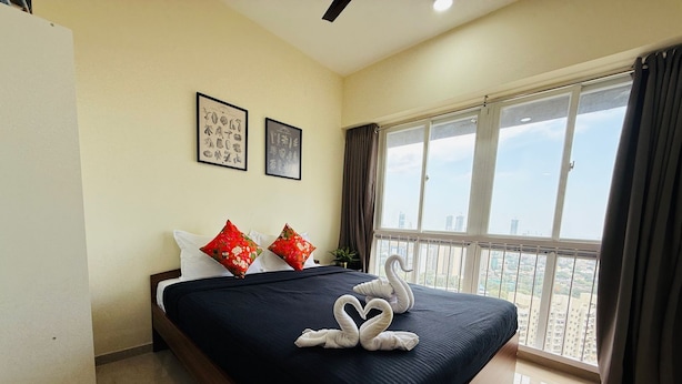 Habitación de hotel con una cama grande, dos almohadas rojas estampadas con flores, una colcha azul oscuro y vistas a un paisaje urbano a través de ventanas amplias.
