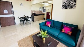 Una sala de estar con un sofá color turquesa, una mesa de centro con una planta, y un control remoto de televisión. Hay una zona de cocina con un microondas y un refrigerador.