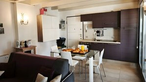 Une cuisine moderne avec des meubles de couleur sombre, un coin repas doté d’une table en verre et un salon avec un canapé foncé.