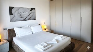Une chambre à coucher avec un grand lit, du linge de lit blanc, un tableau représentant une rose accroché au mur et une armoire aux poignées argentées.