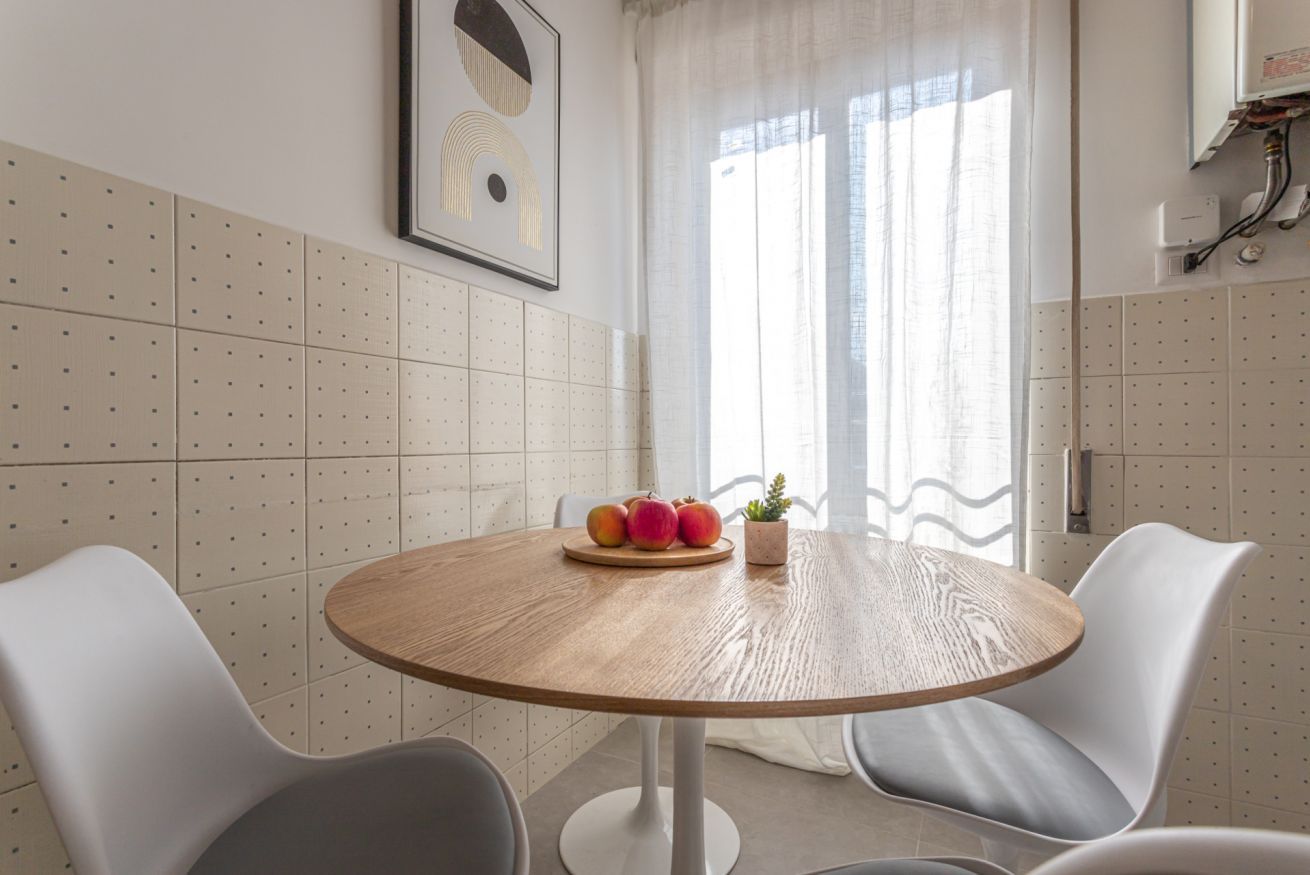 A round wooden table with a fruit centerpiece and a small plant, surrounded by white chairs in a room with a tiled wall and a window with curtains.