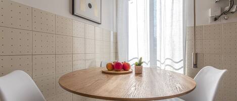 A round wooden table with a fruit centerpiece and a small plant, surrounded by white chairs in a room with a tiled wall and a window with curtains.