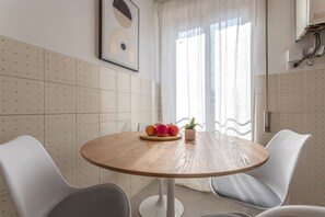 A round wooden table with a fruit centerpiece and a small plant, surrounded by white chairs in a room with a tiled wall and a window with curtains. - LUMINOSO APPARTAMENTO AL PIGNETO (ROMA)