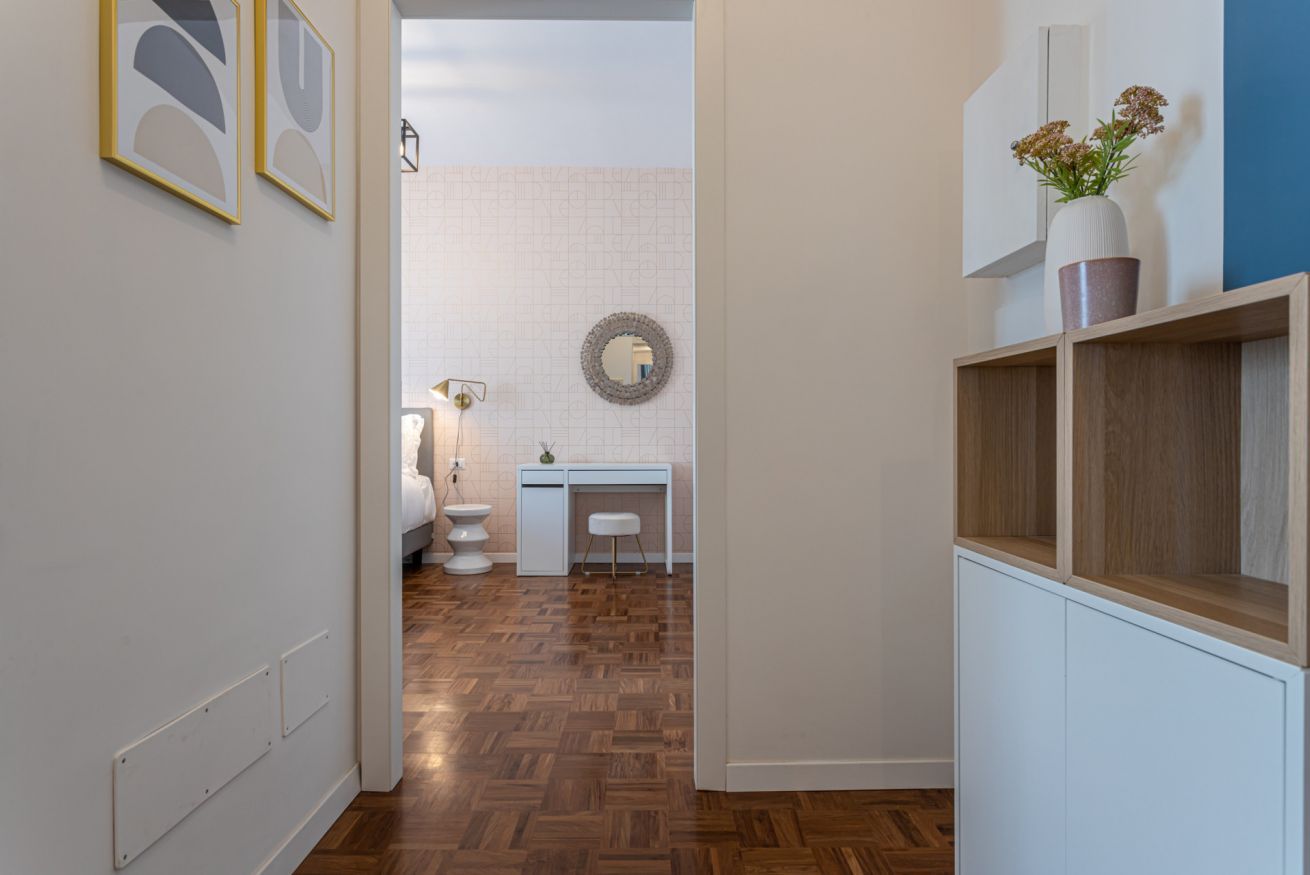 A hallway leading to a bathroom with a toilet, sink, and mirror. There are framed pictures on the wall and a wooden cabinet with a vase on top.