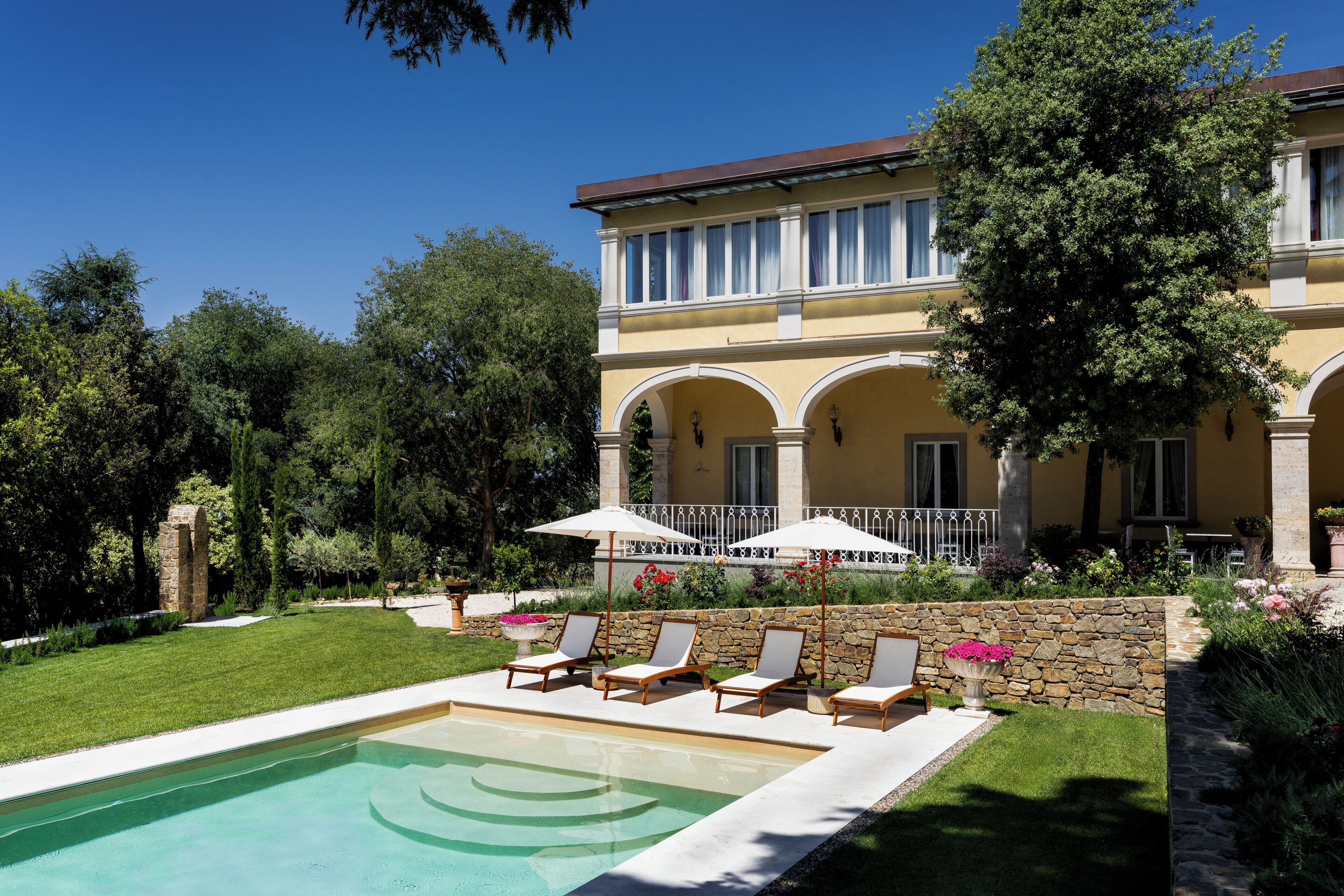 A pool with lounge chairs and umbrellas, a stone wall, and a building with arched windows and a balcony.