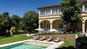 A pool with lounge chairs and umbrellas, a stone wall, and a building with arched windows and a balcony.