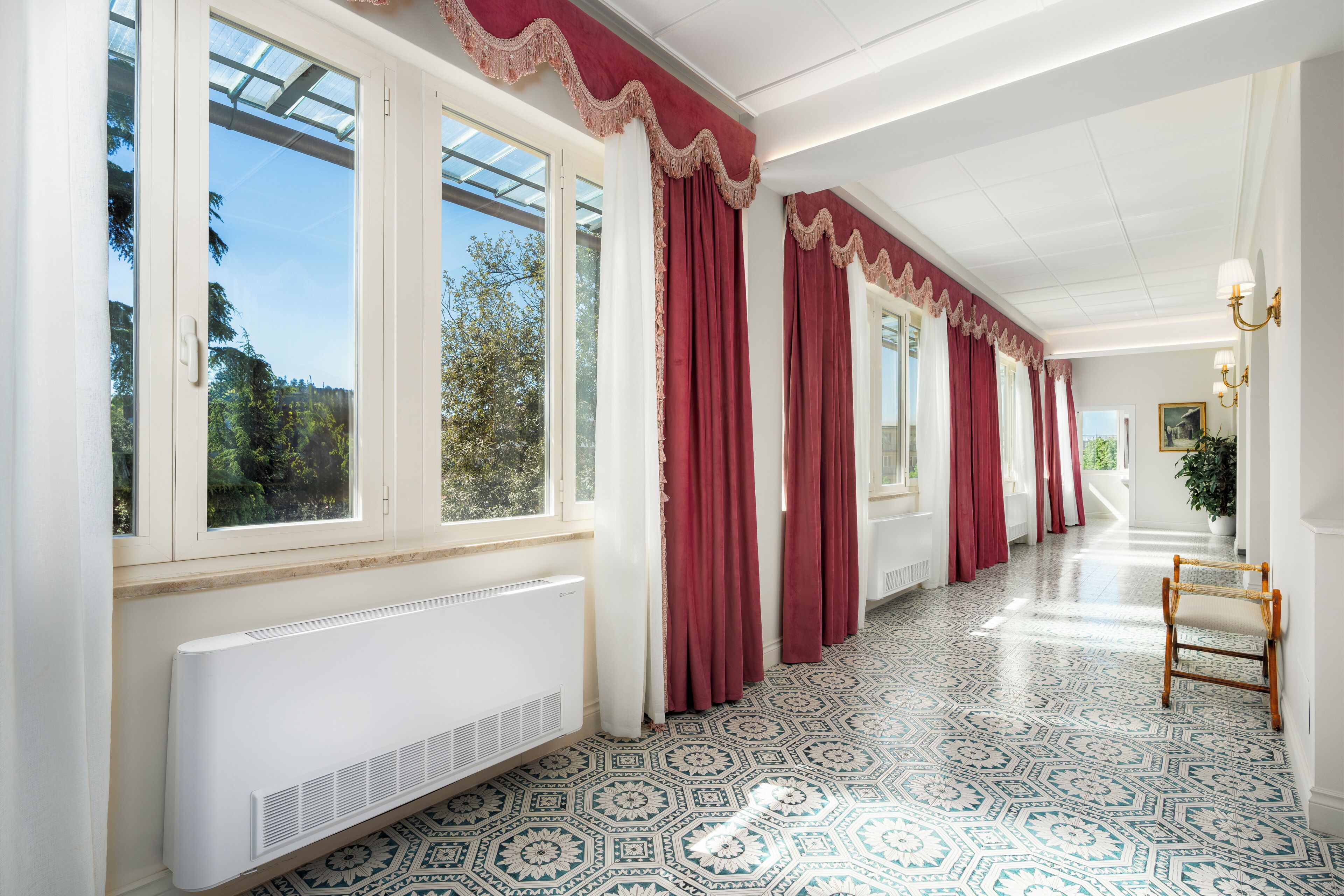 Un pasillo con piso estampado, ventanas amplias con cortinas rojas y un radiador.