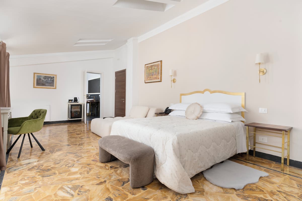 Ein Hotelzimmer mit einem großen Bett, einem grünen Sessel, einer Bank, einem kleinen Tisch und einer Wandlampe.