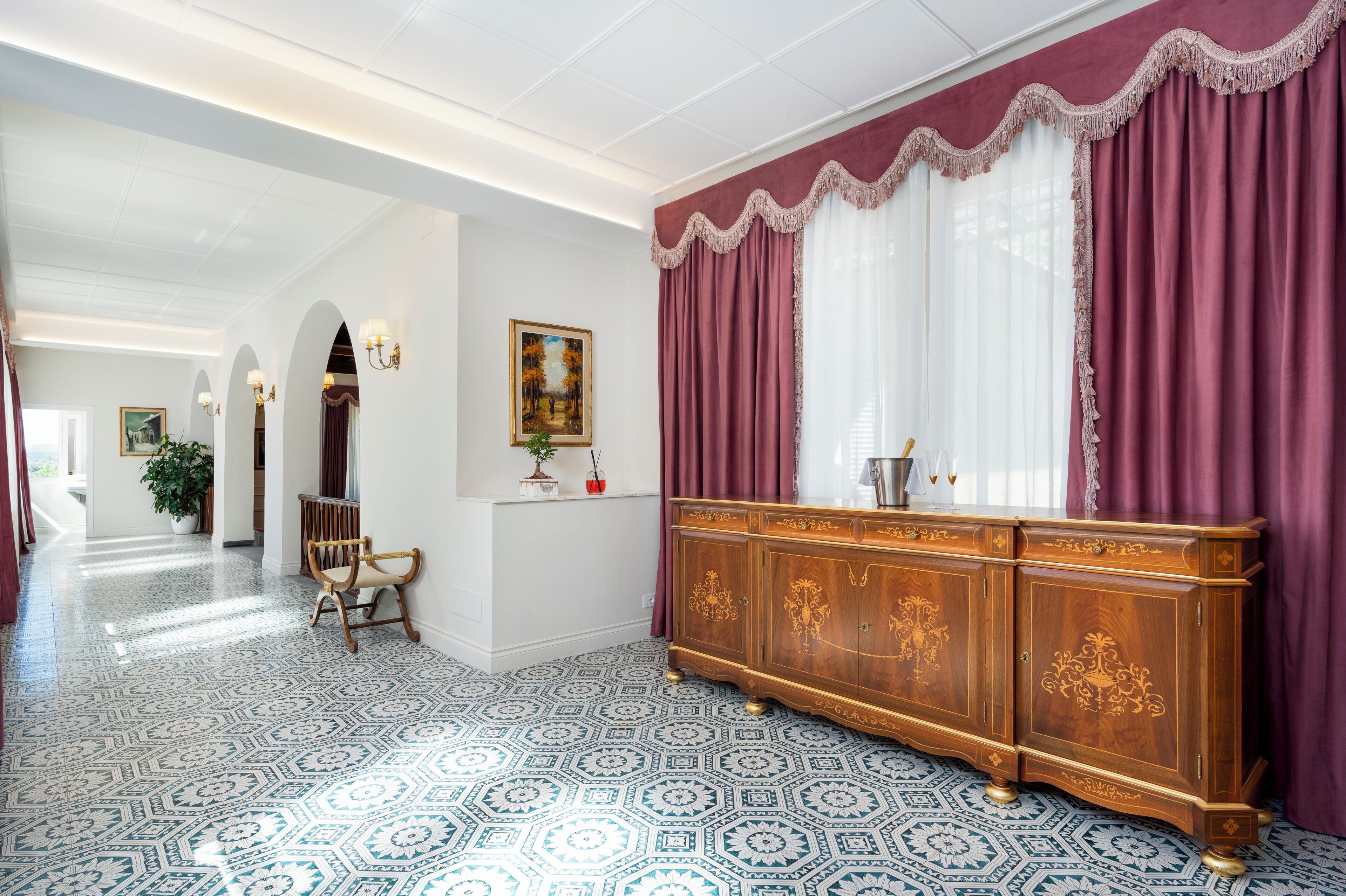 A hallway with patterned flooring, a wooden sideboard, and a large window with curtains.