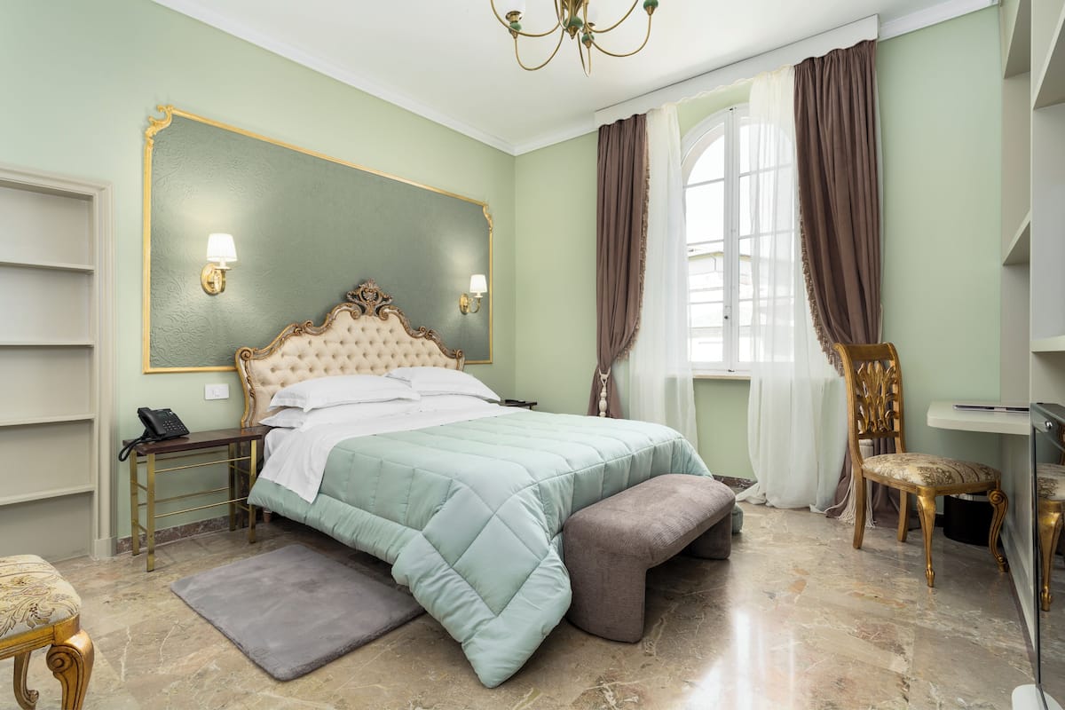 Ein Schlafzimmer mit einem großen Bett, Nachttischen, einem Schreibtisch, einem Sessel und einem Fenster mit Vorhängen.