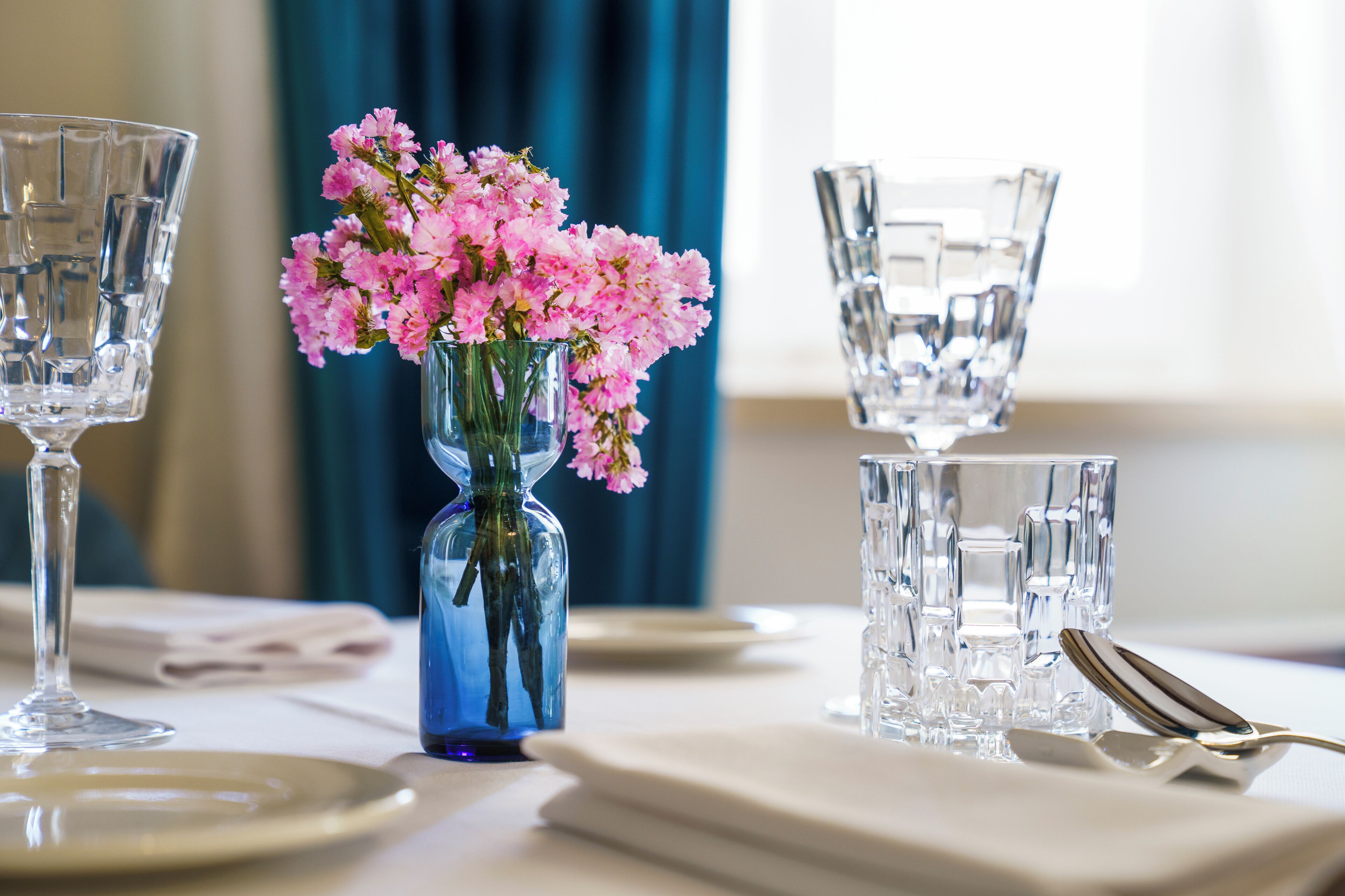 Una mesa de comedor con cristalería transparente, un jarrón azul con flores rosas y una cuchara sobre una servilleta.