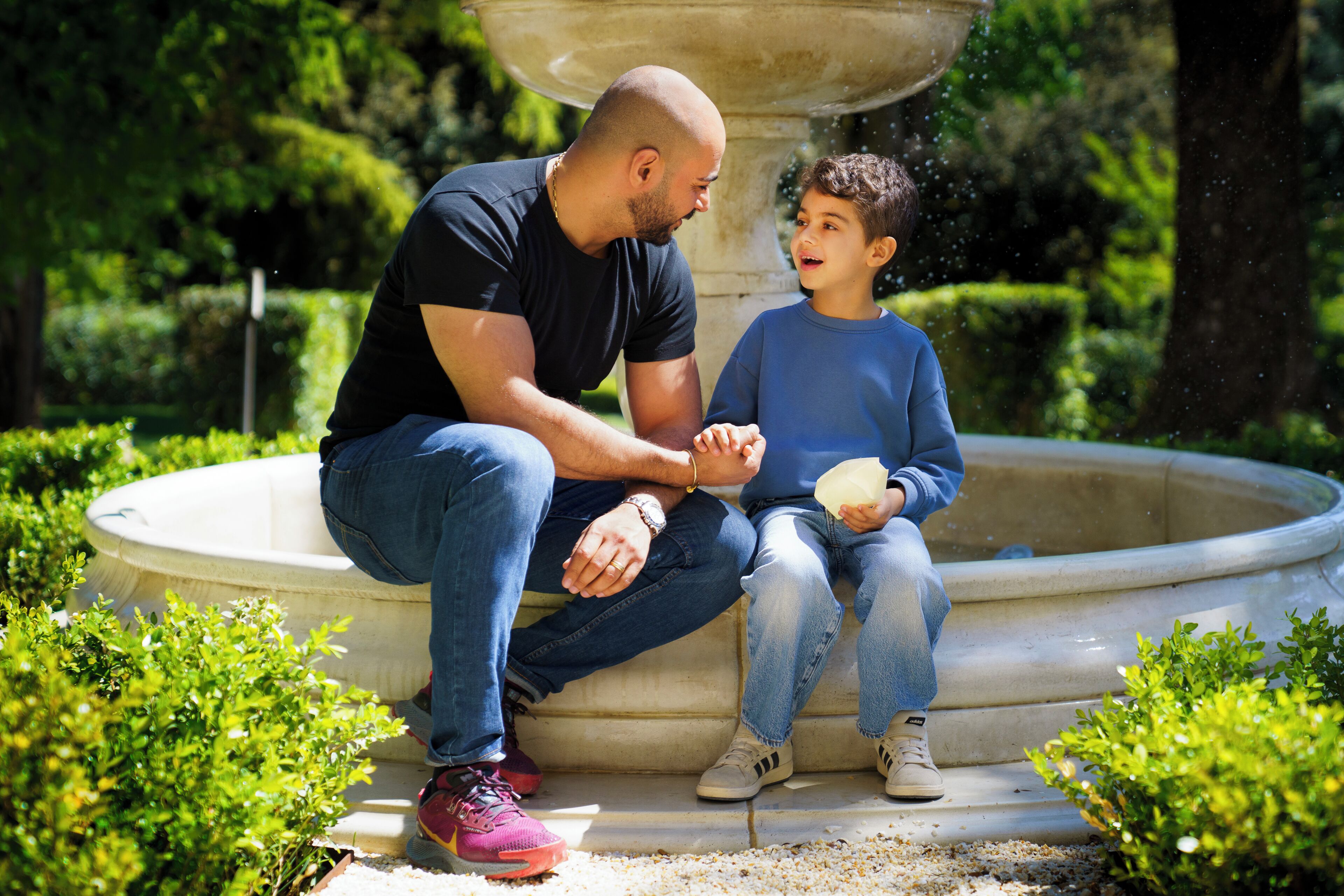 Un homme et un enfant sont assis sur un banc de pierre dans un jardin où se trouve une fontaine.
