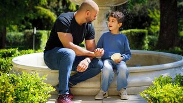 Un hombre y un niño sentados en un banco de piedra en un jardín con una fuente.