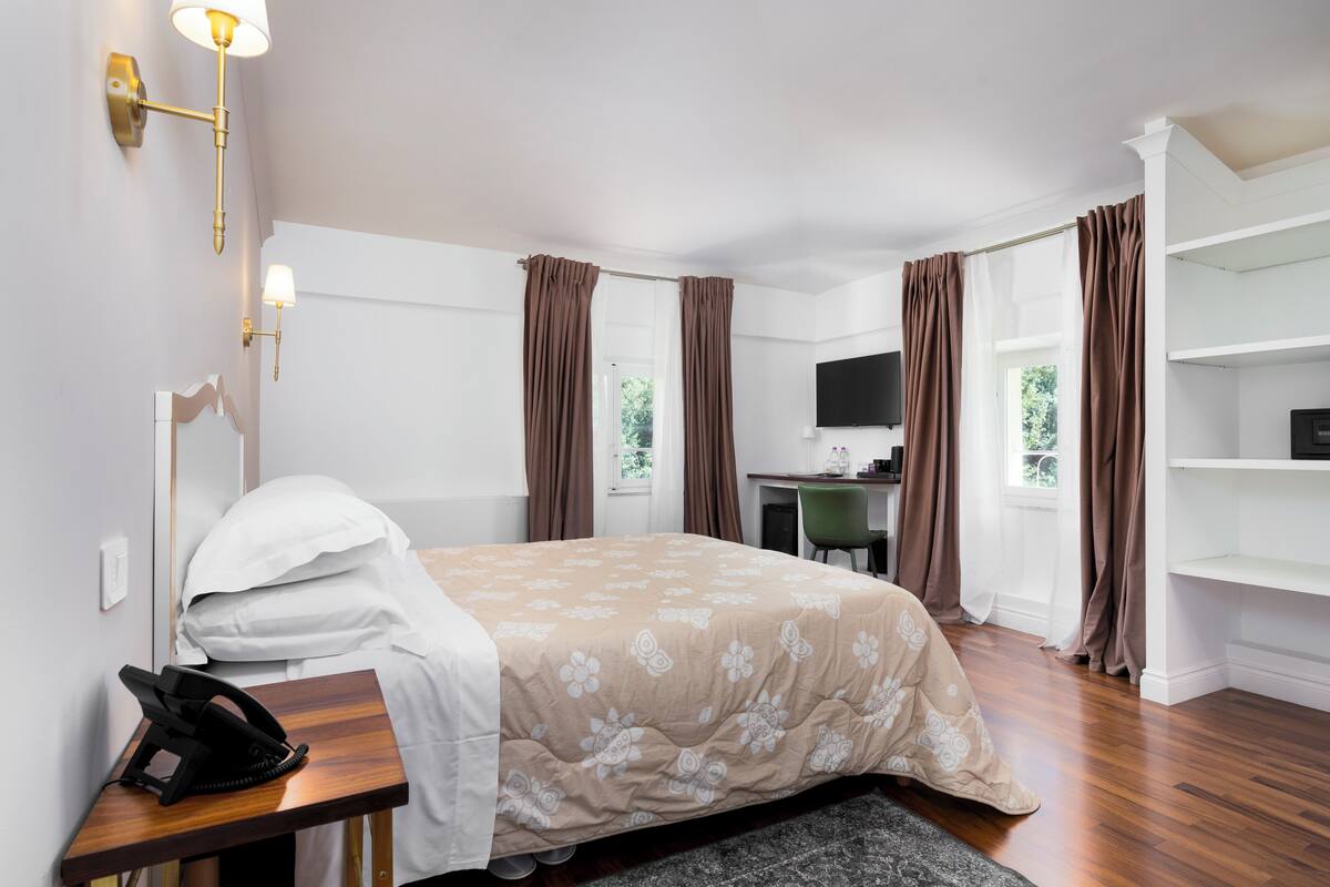 Ein Hotelzimmer mit Bett, Nachttisch, Lampe, Schreibtisch, Stuhl, Fernseher und Vorhängen.