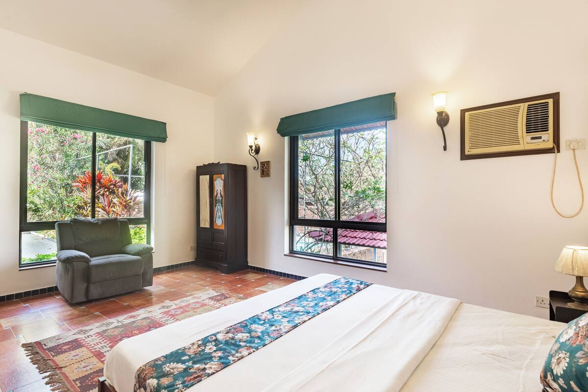 A hotel room with a bed, a chair, a wardrobe, and two windows with curtains.