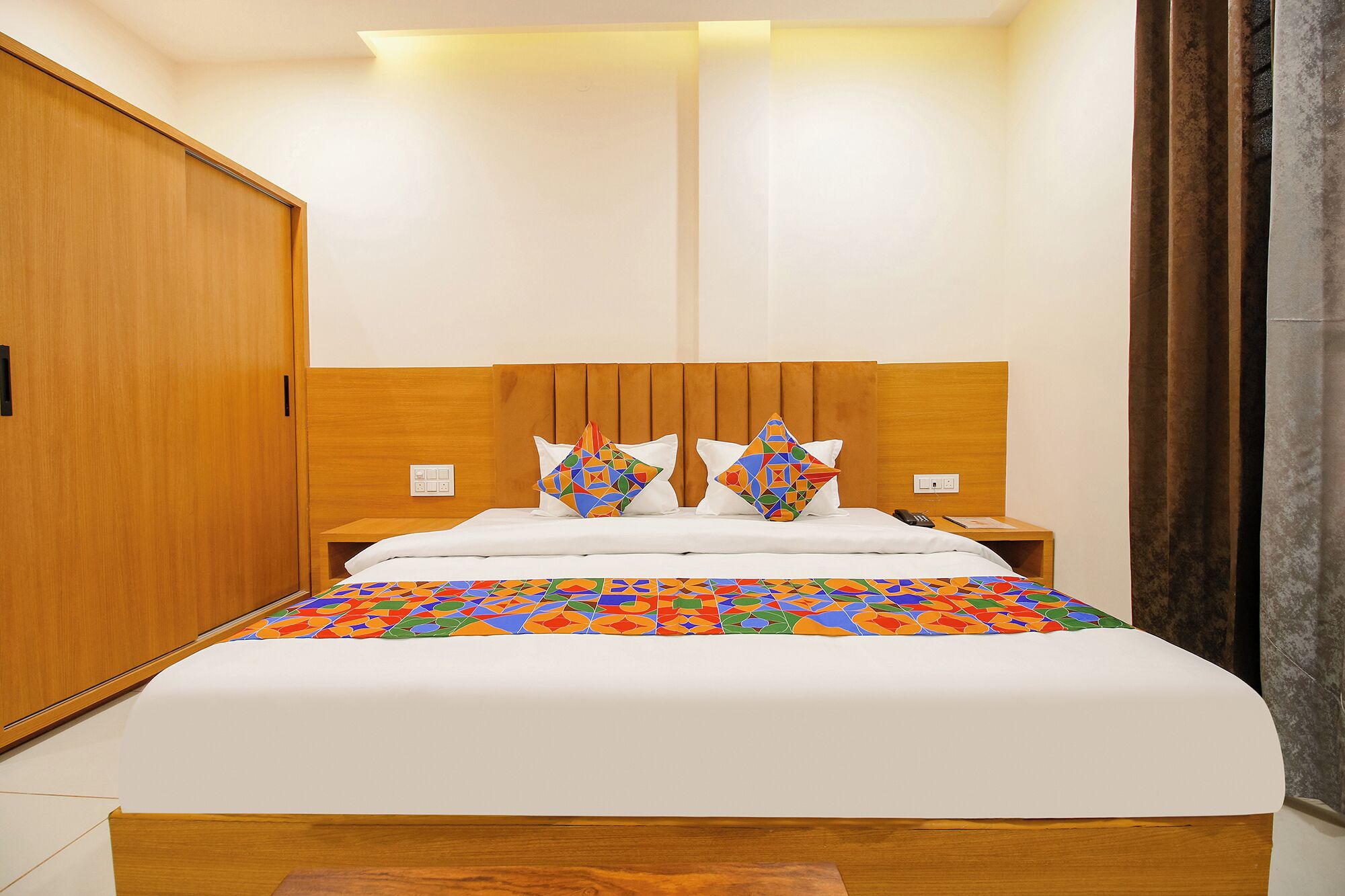 Quarto de hotel com cabeceira de madeira, cama com roupa de cama branca, duas mesas de cabeceira e um edredom colorido com padrão geométrico.