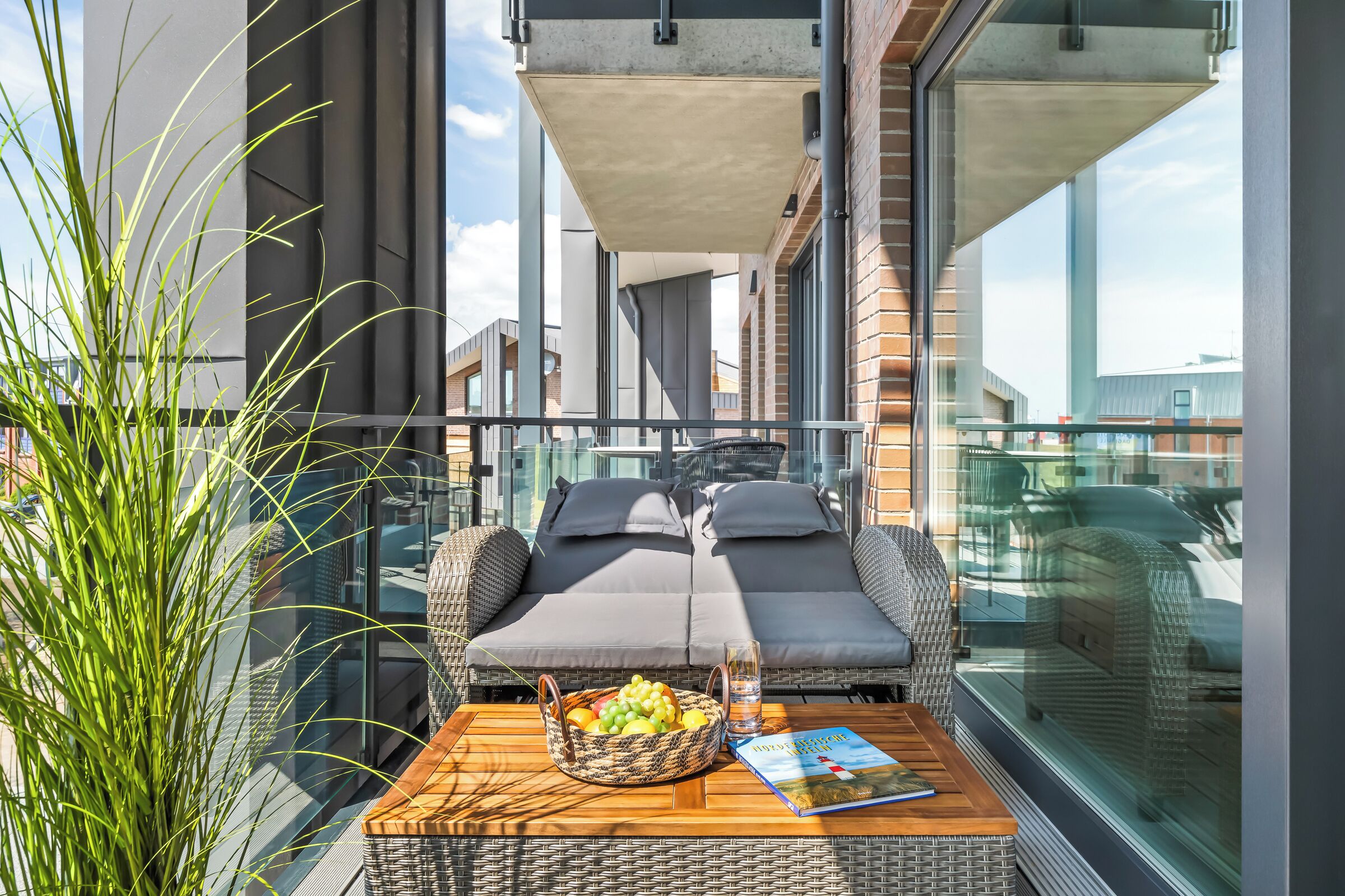 A balcony with a wicker sofa, a wooden table with a fruit basket and a book, and a potted plant.