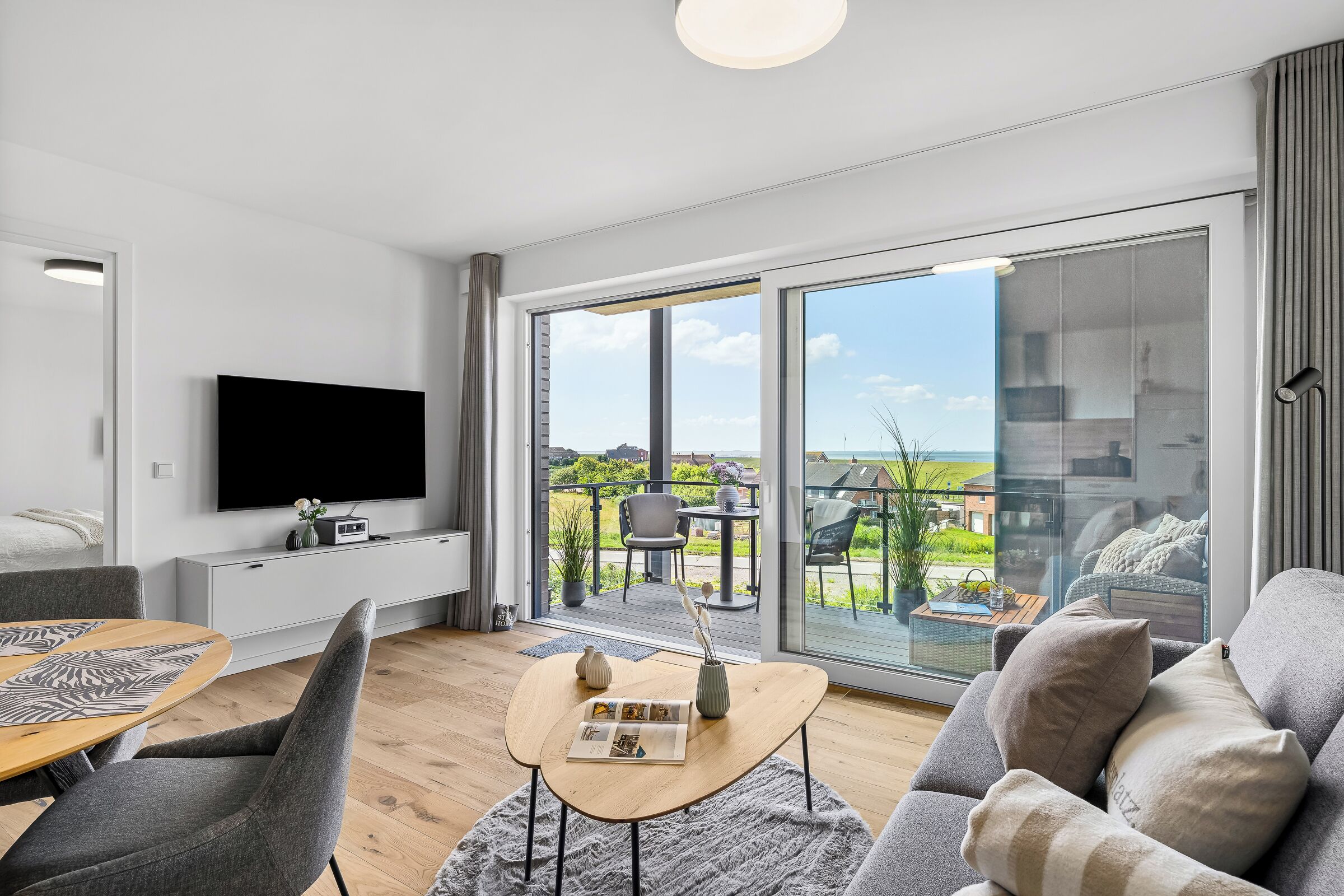 A modern living room with a flat-screen TV, a dining area with a wooden table, and a balcony with a view of the countryside.