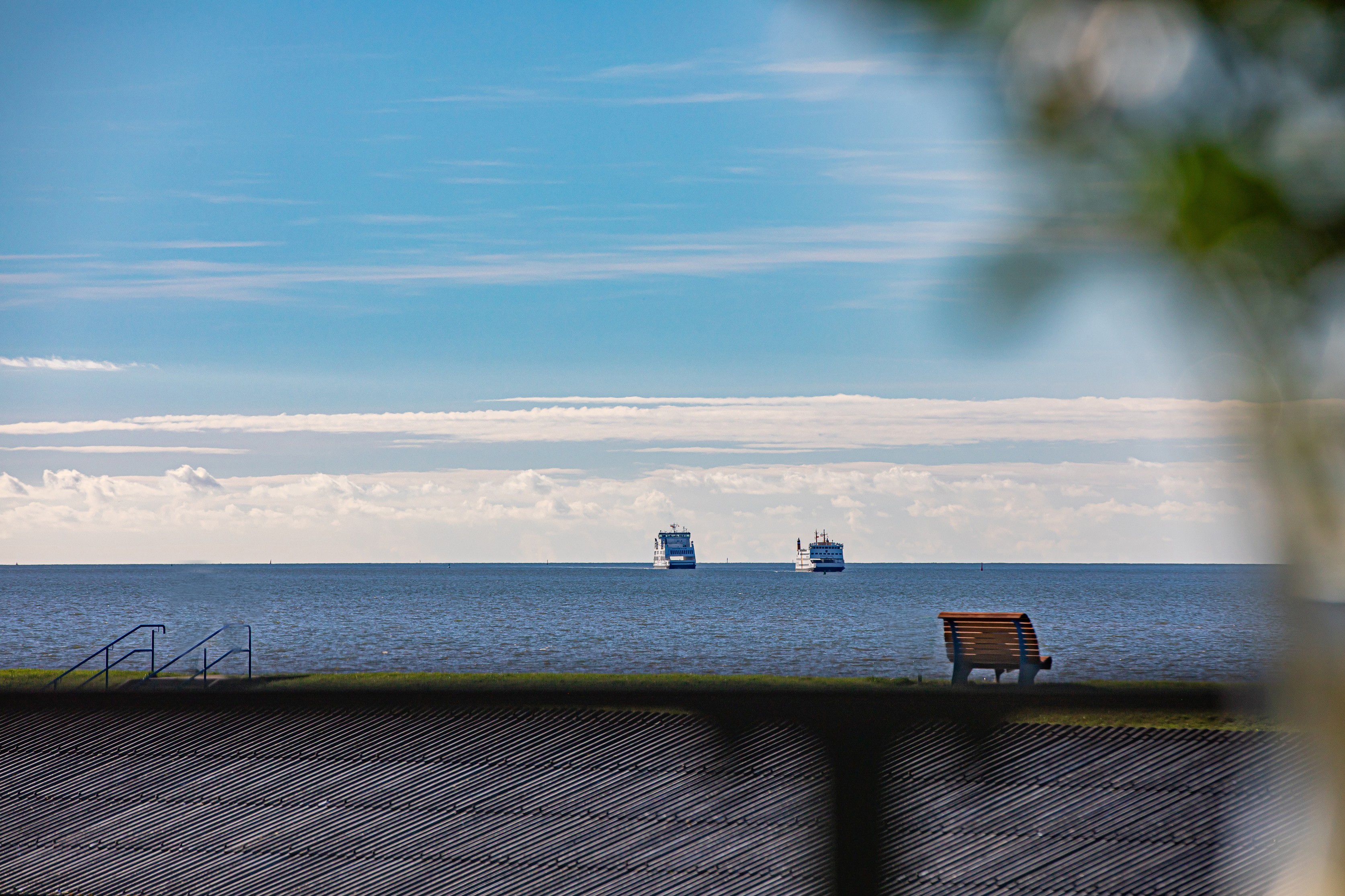 Eine Bank mit Blick auf ein ruhiges Meer, in der Ferne zwei Boote und ein klarer Himmel.