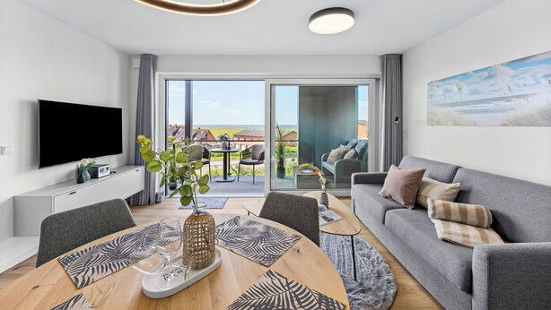 Ett modernt vardagsrum med en platt-TV, en matplats med bord och stolar, en grå soffa och ett stort fönster med utsikt över omgivningen.