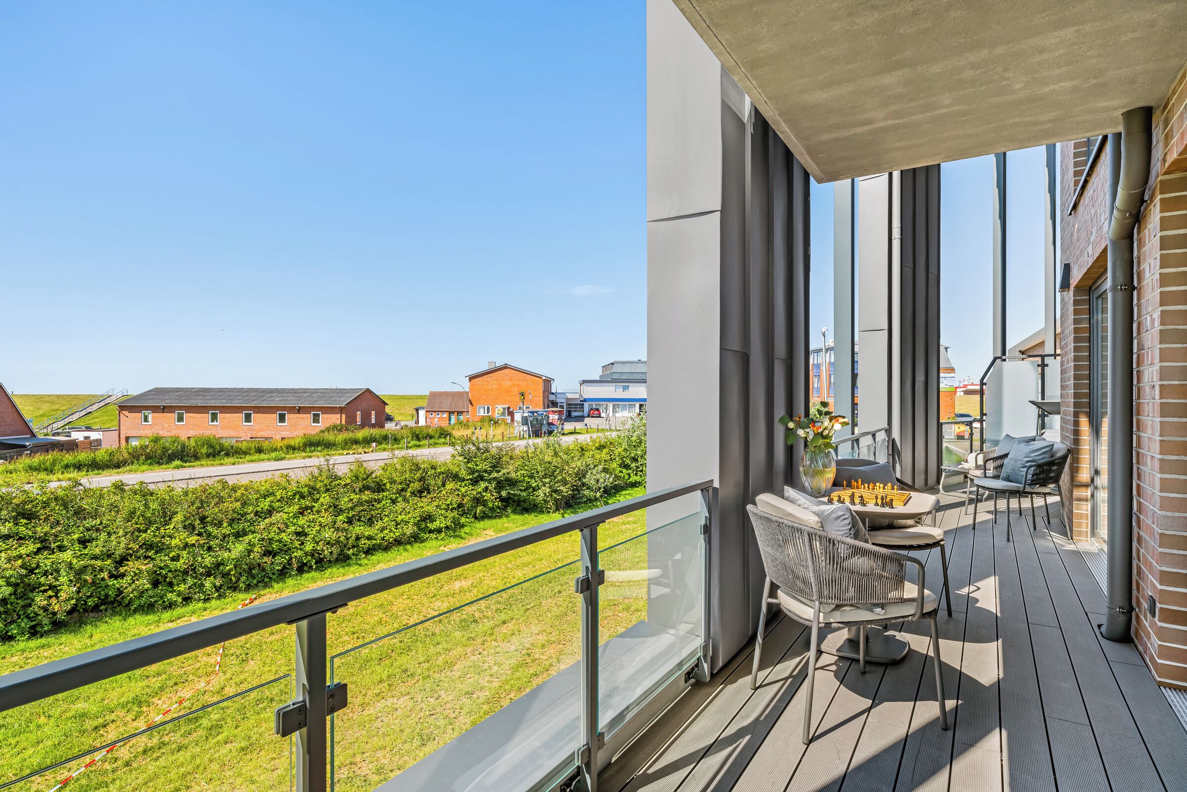 Een balkon met een schaakspel en stoelen, met uitzicht op een gazon en gebouwen.