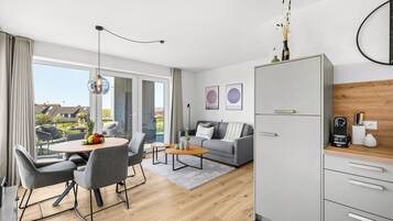 A modern living room with a grey sofa, a dining table with chairs, a coffee machine, and a kitchen area with a white cabinet.