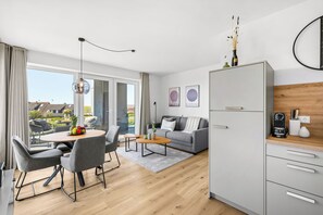 A modern living room with a grey sofa, a dining table with chairs, a coffee machine, and a kitchen area with a white cabinet. - The View B07Johanna - common. Washing machine, balcony, elevator, parking space (Dagebüll)