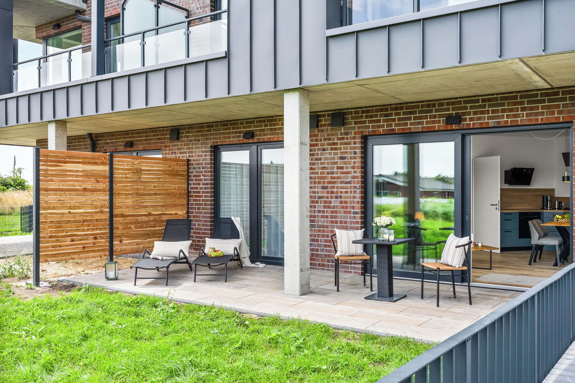 A modern outdoor patio with seating, a wooden fence, and a view into the building.
