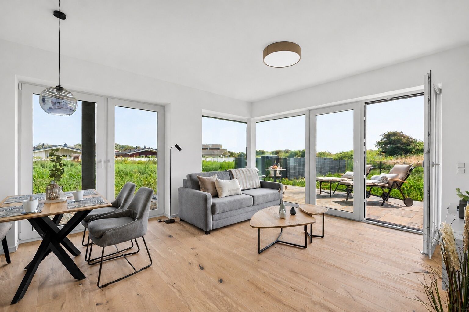 A modern living room with a grey sofa, a dining table with chairs, and a view of a garden through large glass doors.