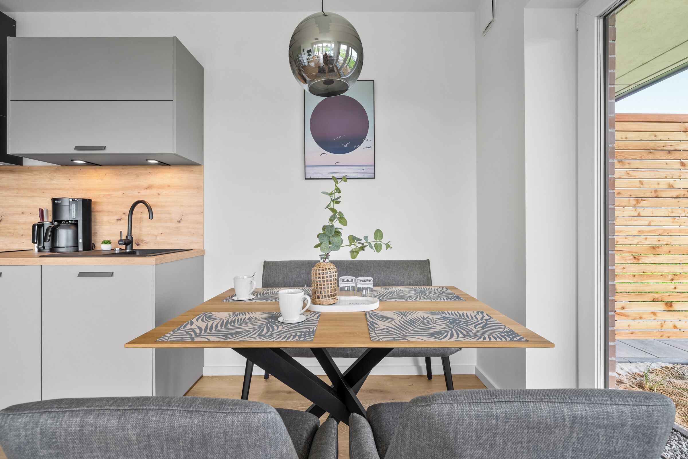 Eine moderne Küche mit Kaffeemaschine, einem hängenden Spiegel und einem gerahmten Kunstwerk über einem Esstisch mit einer Pflanze als Mittelstück.