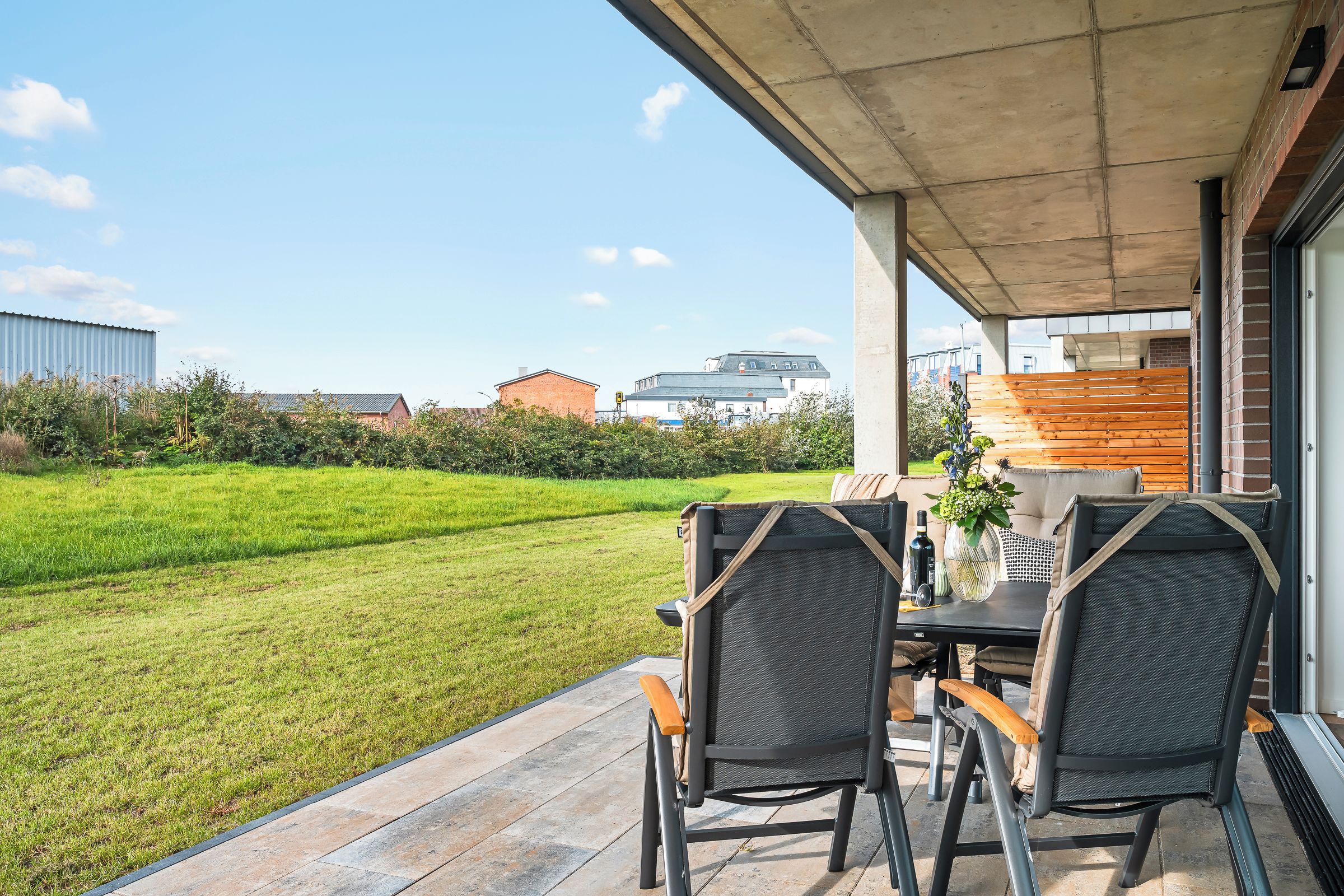 Um pátio externo com mesa e cadeiras, vista para um gramado verde e um edifício ao fundo.