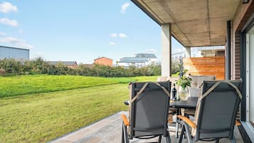 Um pátio externo com mesa e cadeiras, vista para um gramado verde e um edifício ao fundo.