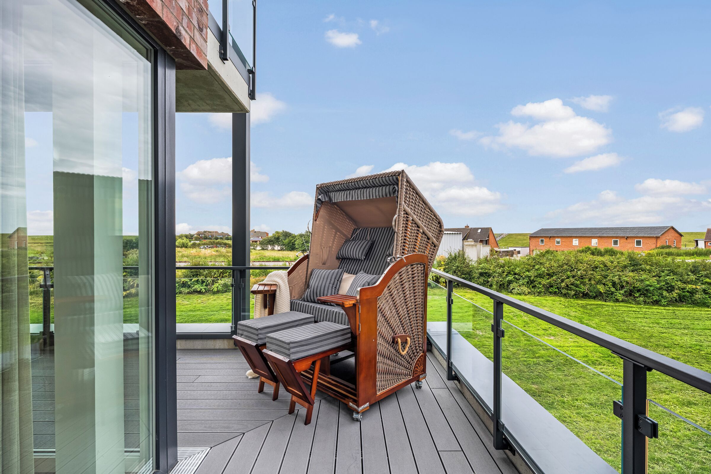 A balcony with a wicker chair and a small table, overlooking a grassy area and buildings.