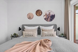 Ein Schlafzimmer mit grau gestopfter Bettdecke, zwei Kissen mit dem Aufdruck 'time to relax', einer Strickdecke und Wandbildern mit Pflanzenmotiven.