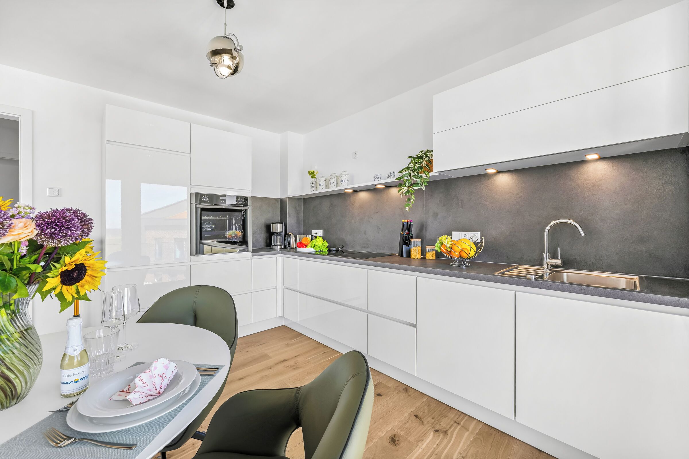 A modern kitchen with white cabinets, a built-in oven, and a sink. There is a dining table with chairs and a vase of flowers.