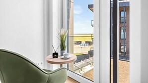Ein modernes Zimmer mit einem grünen Sessel, einem kleinen Tisch mit einer Pflanze und einer Tasse, und Blick auf ein Gebäude und eine Straße.