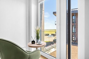 A modern room with a green chair, a small table with a plant and a cup, and a view of a building and a road outside. - The View B09 Austernfischer - Sea view, balcony, shared. Sauna, parking space (Dagebüll)