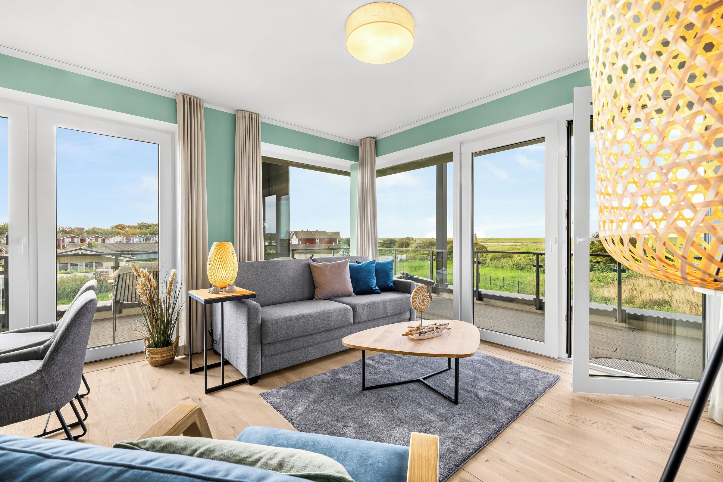 Ein geräumiges Wohnzimmer mit grauem Sofa, einem Couchtisch und Blick auf eine grüne Landschaft durch große Fenster.