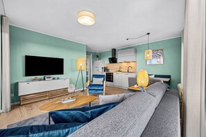 A modern living room with a flat-screen TV, a sofa, a coffee table, and a kitchen area with cabinets and a stove. - The View C06 Strandflieder - 1st floor, shared sauna, parking (Dagebüll)