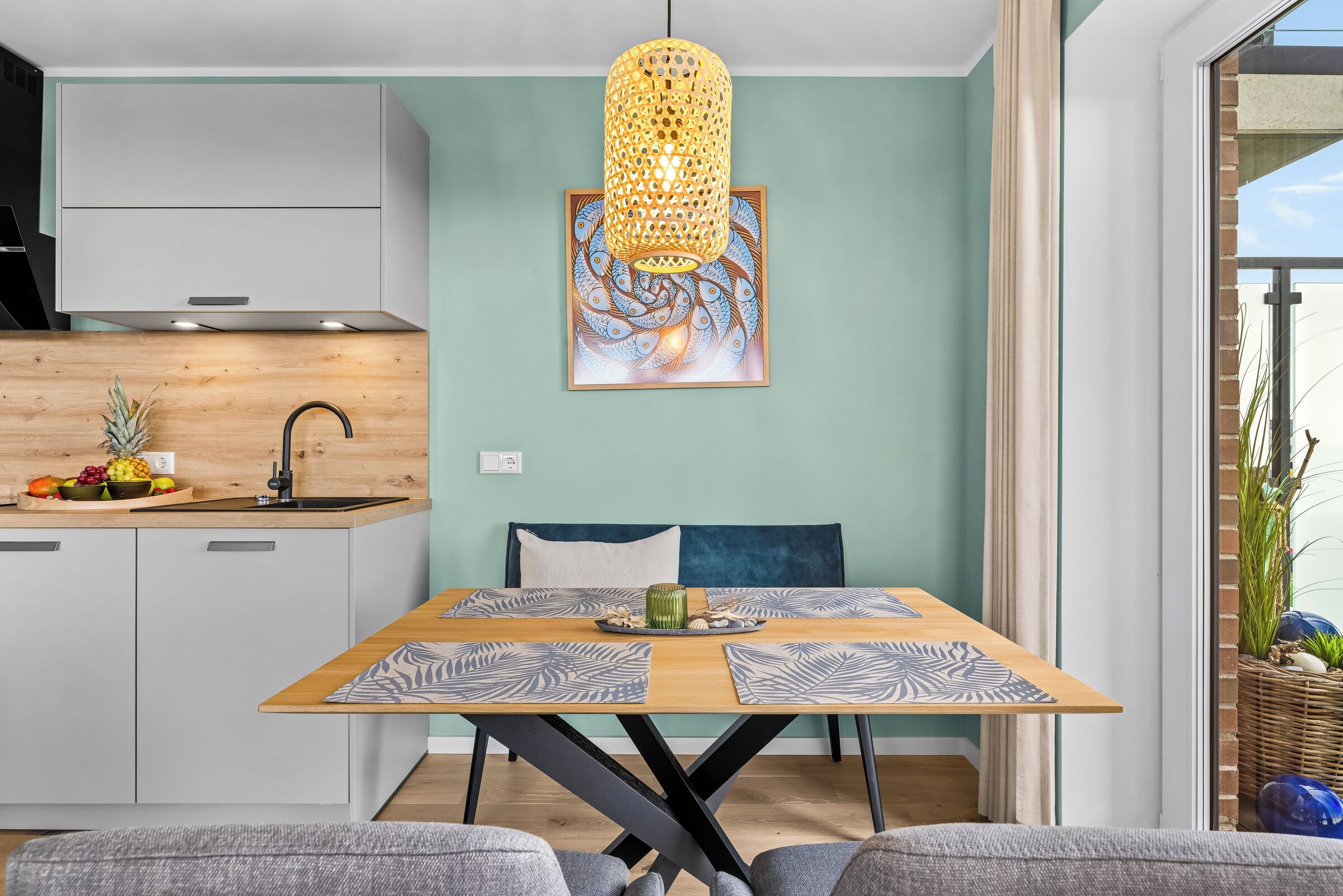 A modern kitchen with a dining area, featuring a wooden table with patterned placemats, a hanging light fixture, and a wall-mounted painting.