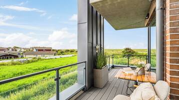 Een balkon met uitzicht op een groen veld, een rieten plantenbak en een tafel met kussen.