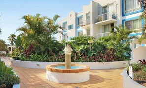 Un patio de hotel con una fuente central, rodeado de abundante vegetación y edificios de varios pisos con balcones.