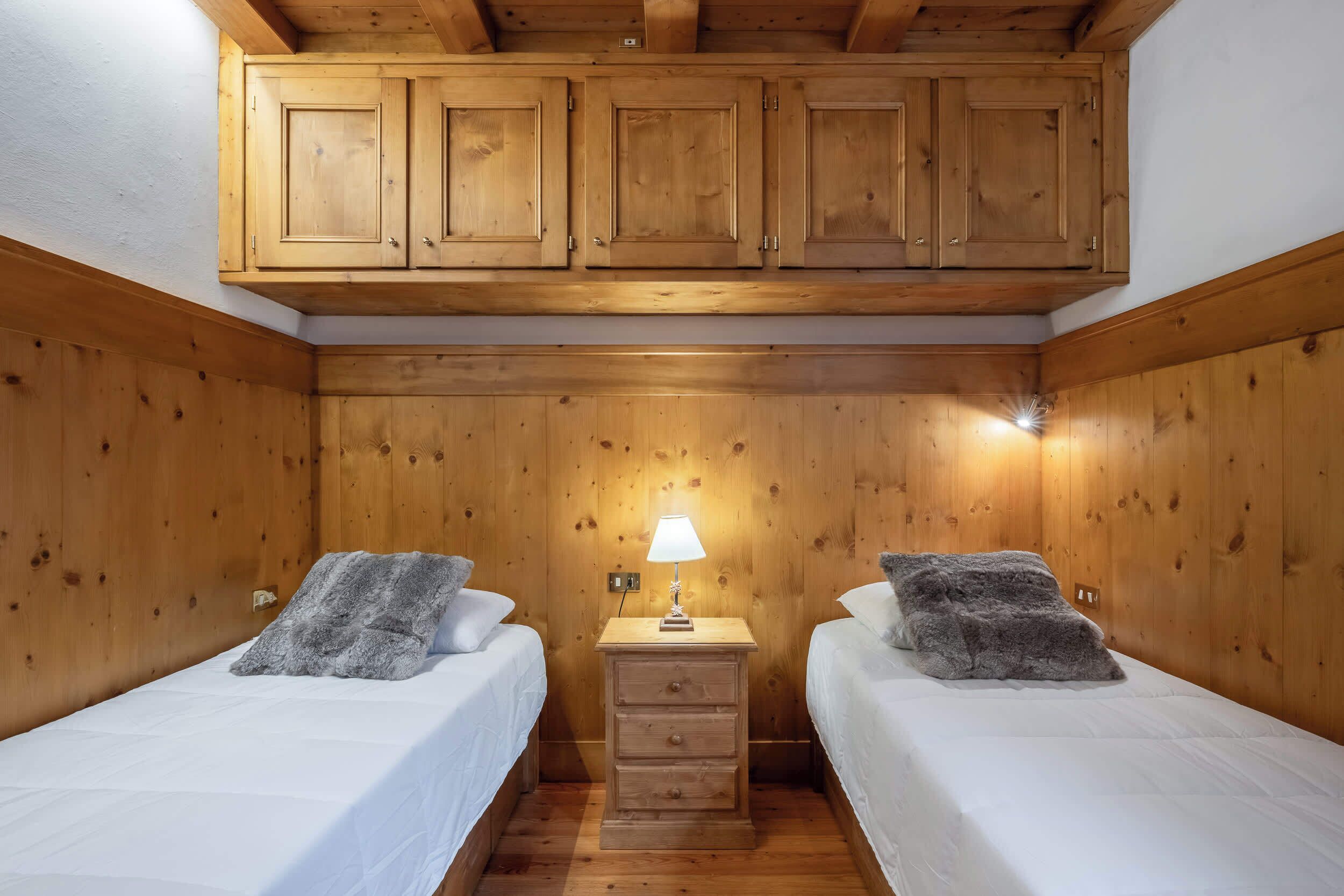 A small, cozy room with wooden walls and a ceiling, featuring two beds with white bedding and fluffy gray pillows, a small wooden nightstand with a lamp, and a wooden cabinet above the beds.