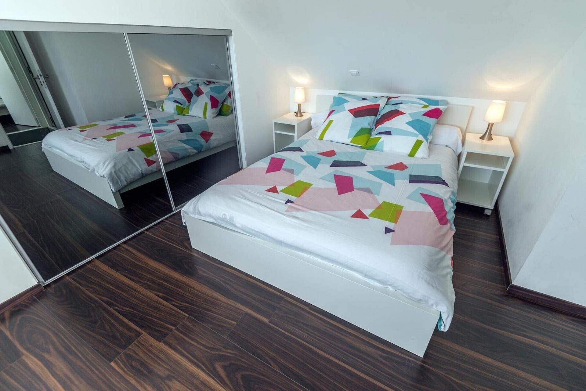 Ein Schlafzimmer mit einem raumhohen Spiegelkleiderschrank, einem Bett mit geometrischem Muster auf der Bettdecke, Nachttischlampen und einem dunklen Holzboden.