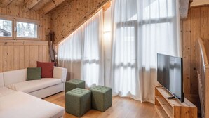 A wooden cabin interior with a sofa, ottomans, a TV on a wooden stand, and large windows with sheer curtains. - Brand new apartment in San Vito di Cadore (San Vito di Cadore)