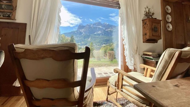 A cozy room with a wooden chair, a table, and a view of mountains through a window with curtains. - Charming ground floor flat with mountain views (Cortina d'Ampezzo)